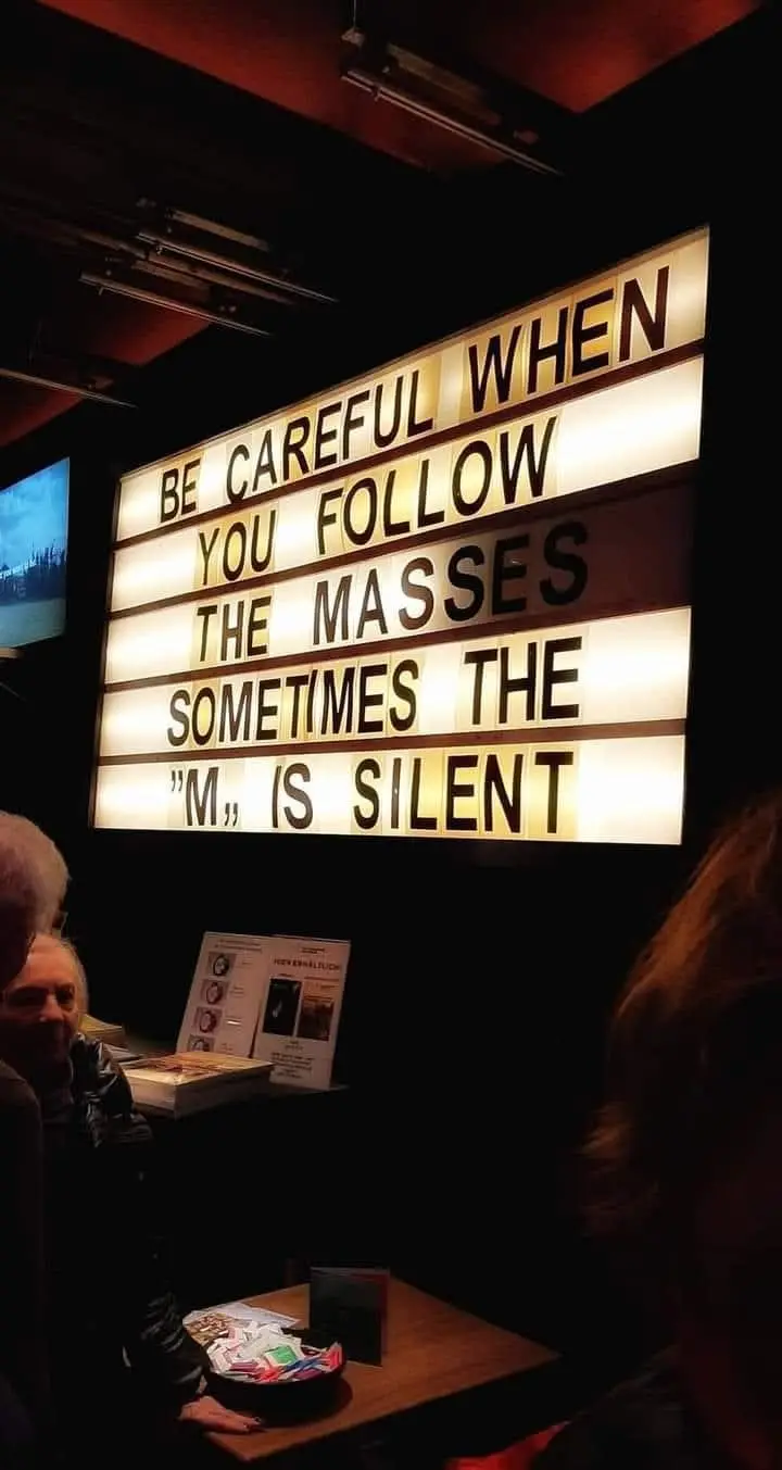 A lit-up marquee sign in a dimly lit venue displays the text 'BE CAREFUL WHEN YOU FOLLOW THE MASSES SOMETIMES THE "M" IS…