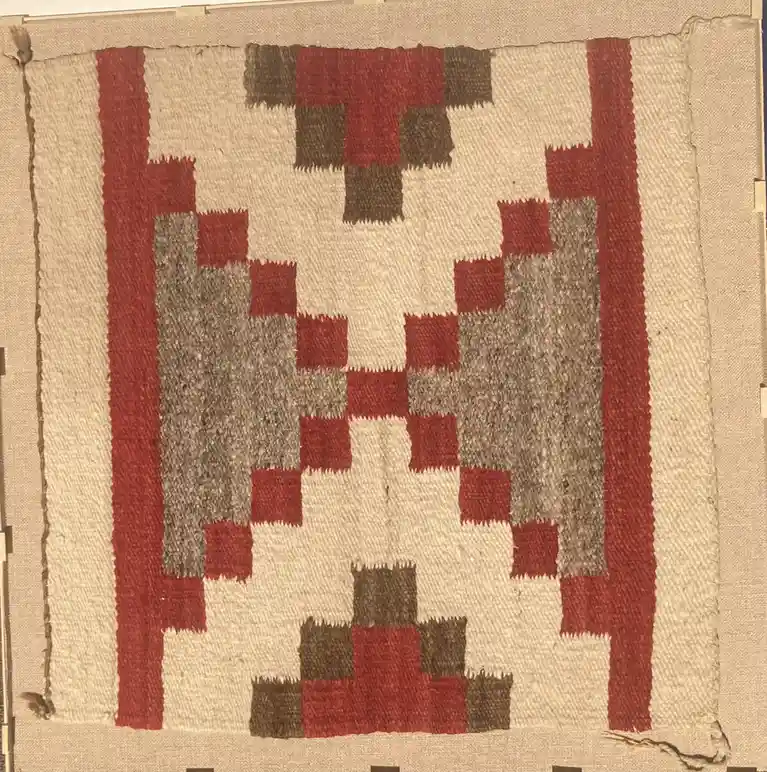 a close-up photograph of a traditional woven textile, likely a Navajo rug, featuring a geometric pattern in red, beige, and…