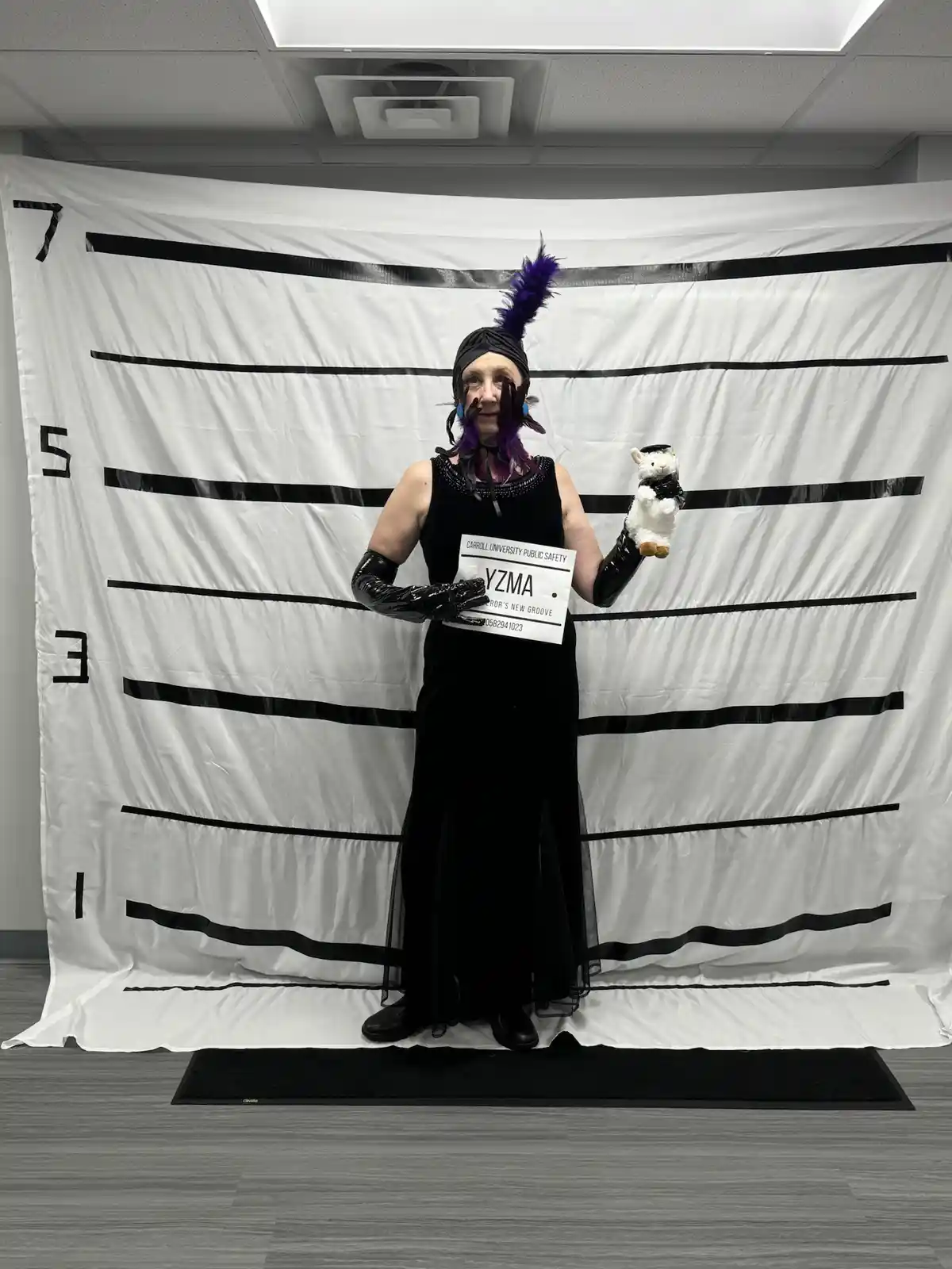 A person dressed in a black flapper-style gown with a purple feather headpiece and black gloves stands in front of a white…
