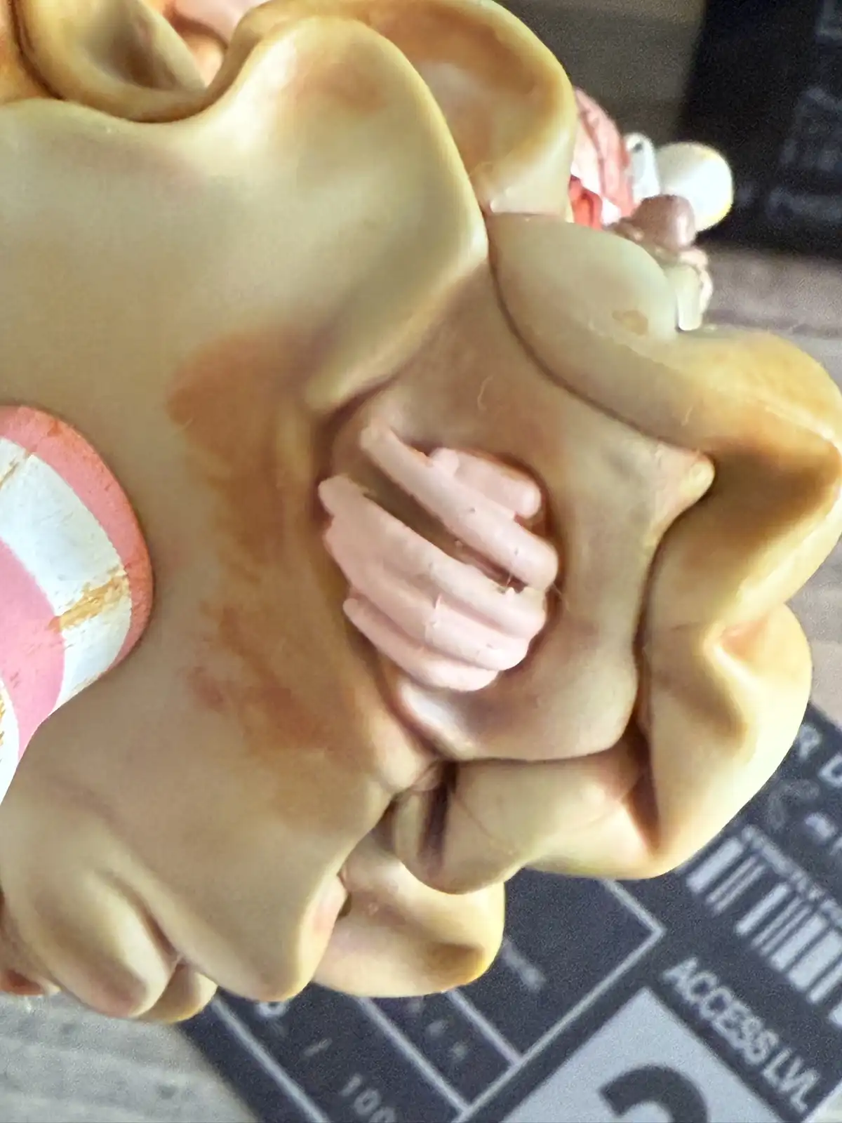 A close-up of a beige, sculpted figurine resembling a stylized flower or abstract face, with a small pink hand-shaped detail…