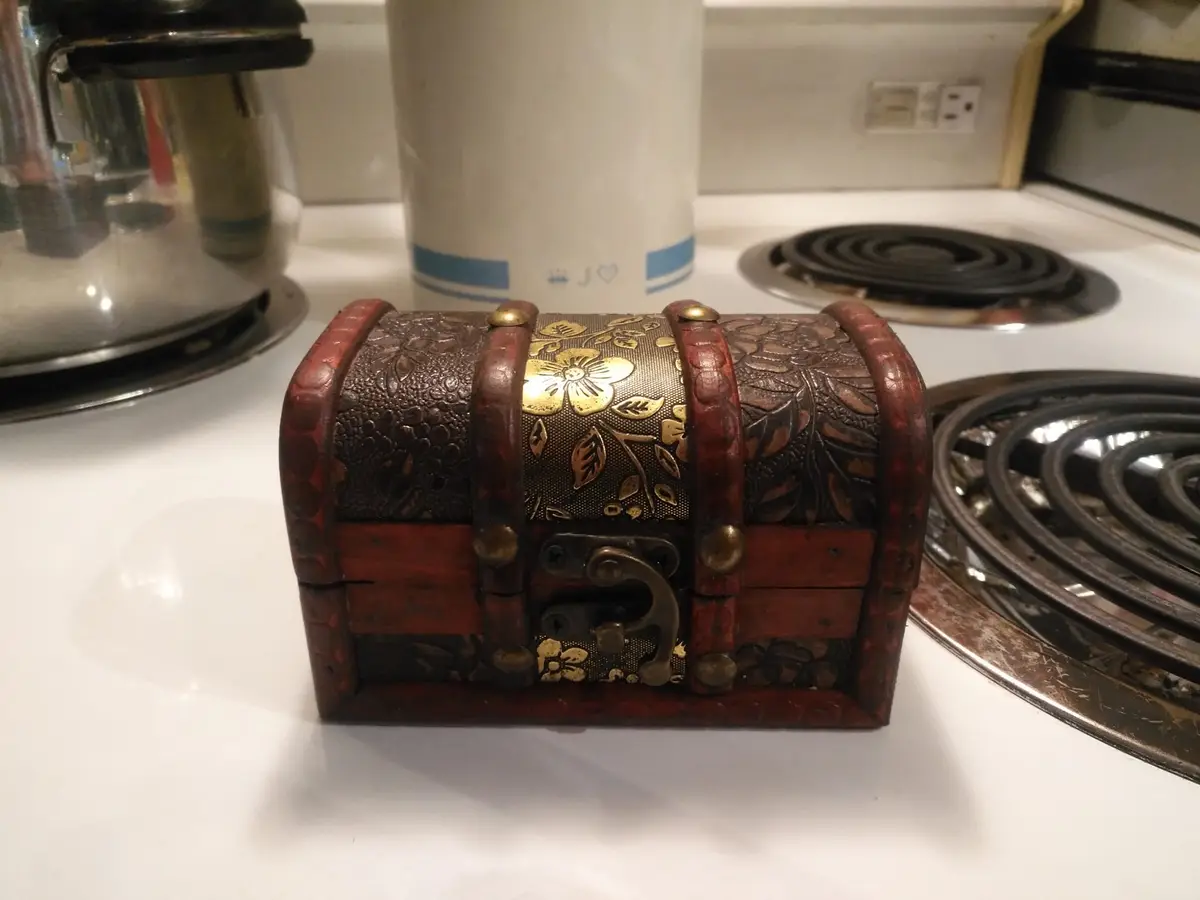 A small, ornate wooden treasure chest with gold floral inlays and brass hardware sits on a white kitchen stovetop…