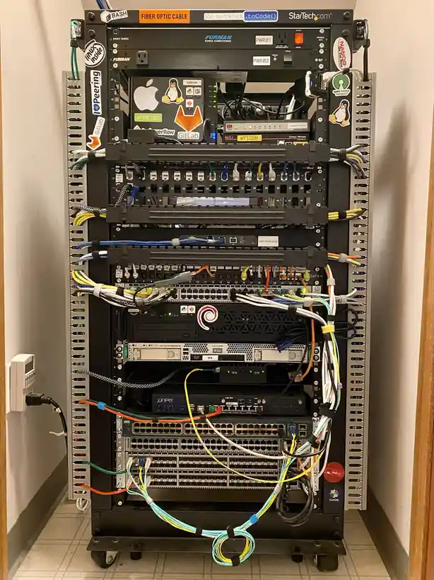 A tall, black server rack filled with networking equipment, cables, and stickers, including Linux, GitLab, and Juniper…