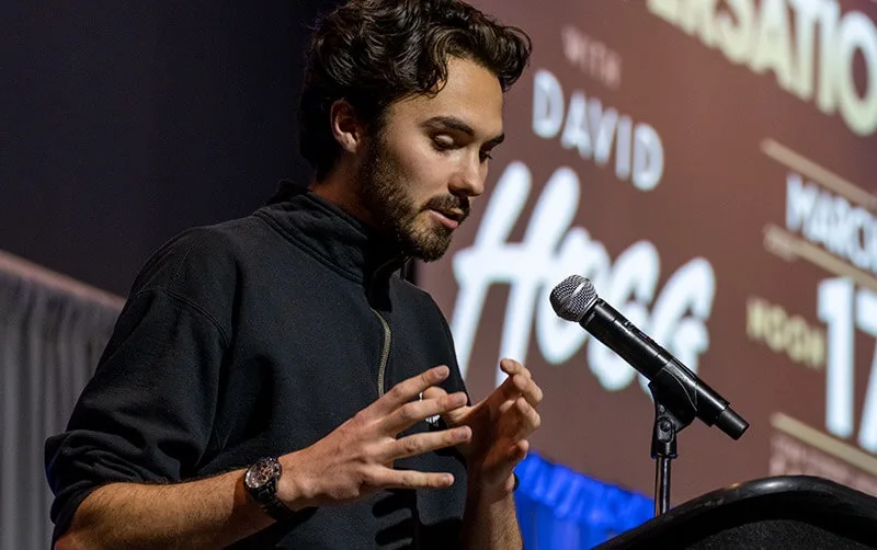 A man with dark hair and a beard, wearing a black zip-neck sweatshirt and a watch, stands at a podium speaking into a…