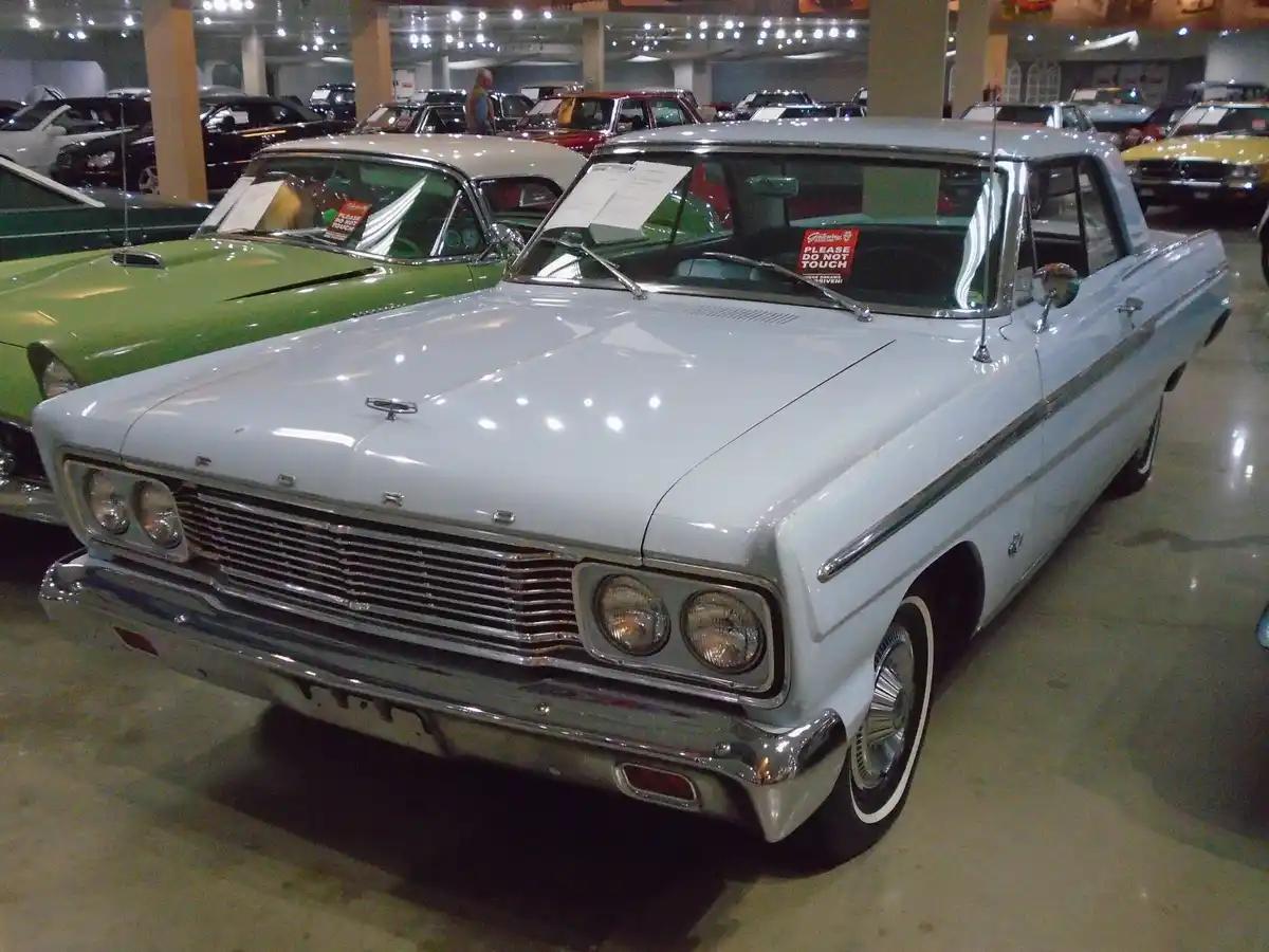A light blue classic Ford car is displayed in a museum or showroom, surrounded by other vintage cars under bright lights. A…