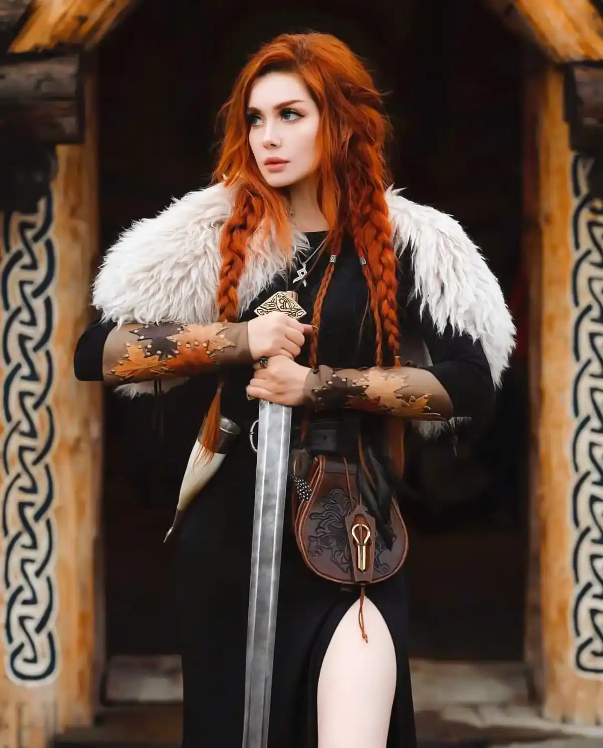 A woman with long red braided hair stands in front of a wooden structure with Norse-style carvings, wearing a black dress…