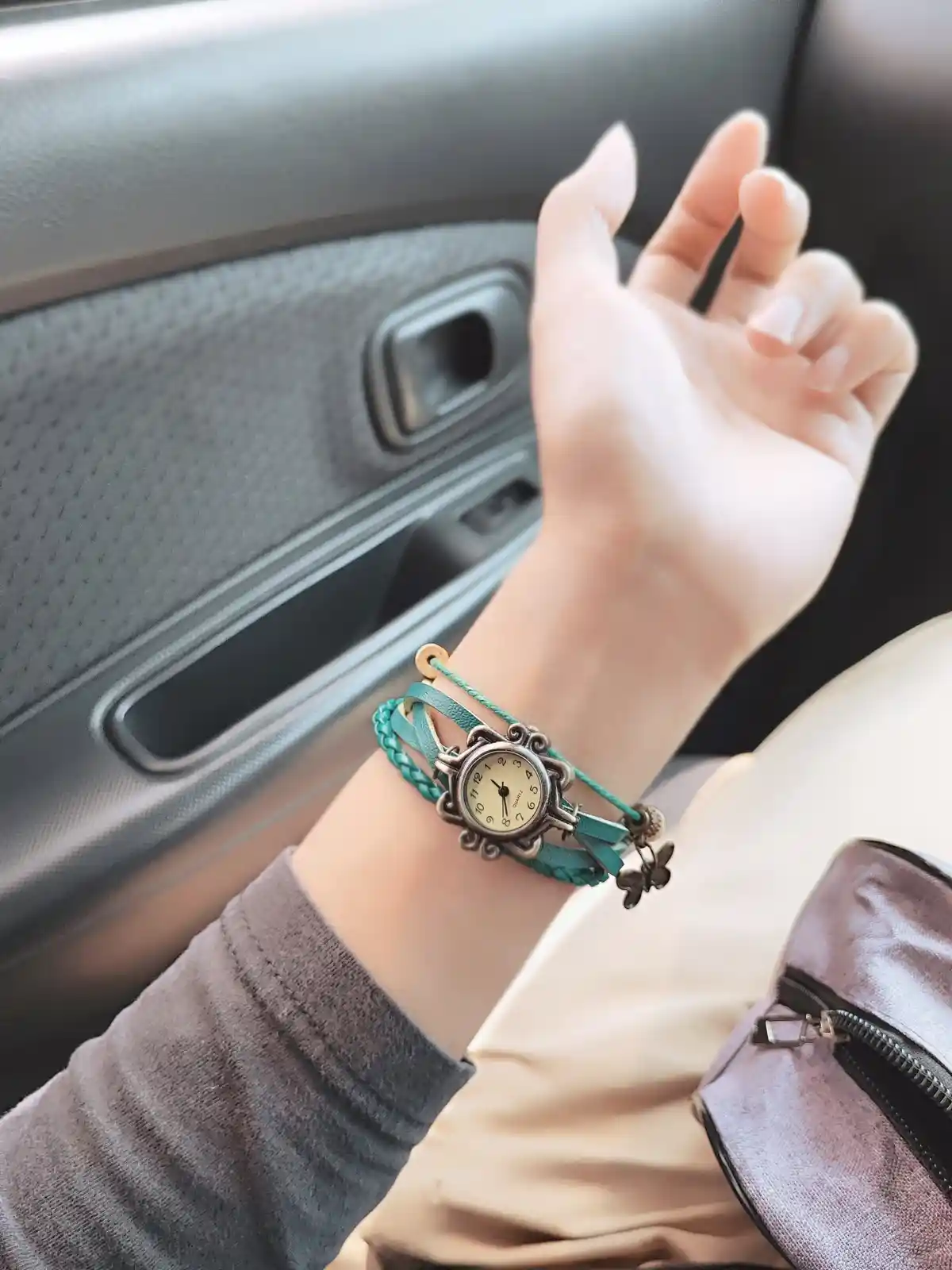 A person's hand rests on a car seat, wearing a turquoise braided bracelet watch with a butterfly charm. The watch face shows…