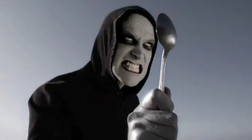 A person in a black hooded robe with pale makeup and an angry expression holds up a metal spoon. The image has the text 'i'm…