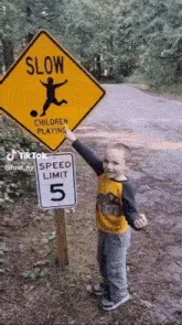 A young boy stands next to a yellow 'SLOW CHILDREN PLAYING' road sign with a speed limit of 5, then runs away as a lion…