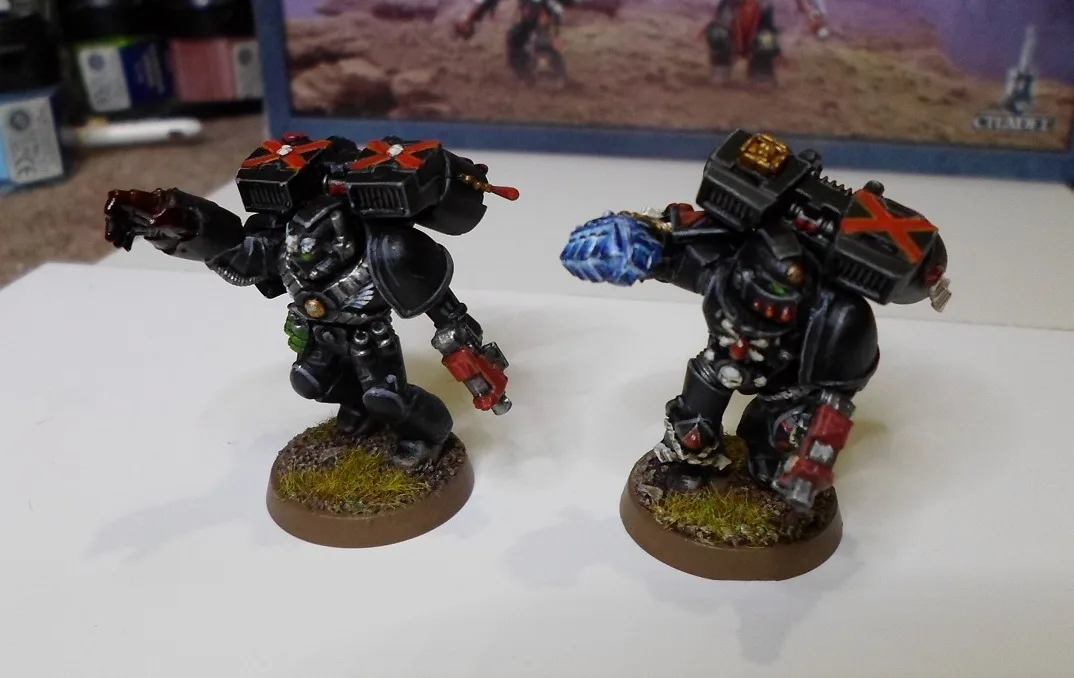 Two painted miniature figures in black power armor with red X markings on their backs stand side by side on circular bases…
