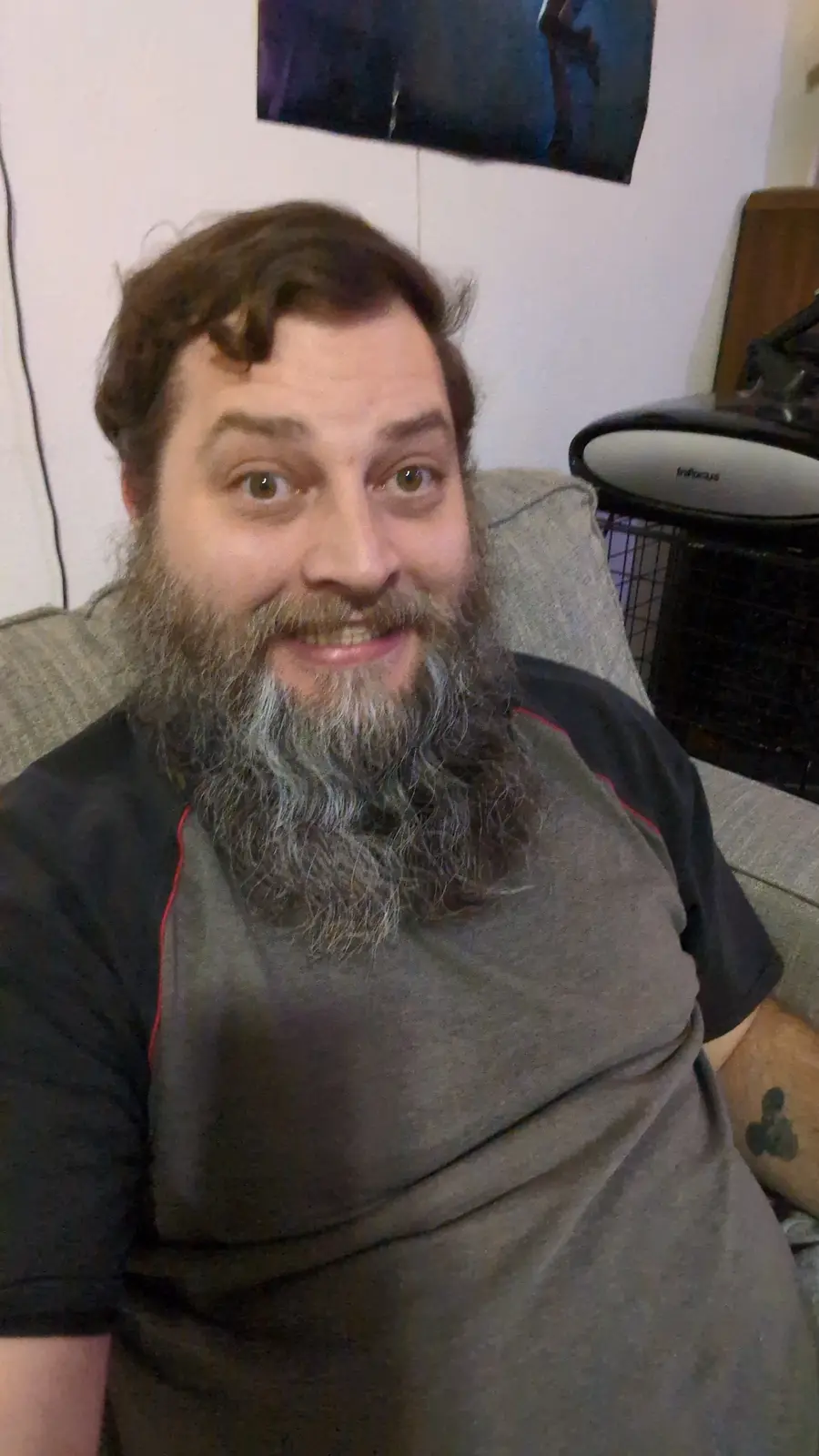 A man with a long, graying beard and brown hair smiles at the camera while sitting on a gray couch. He wears a gray and…