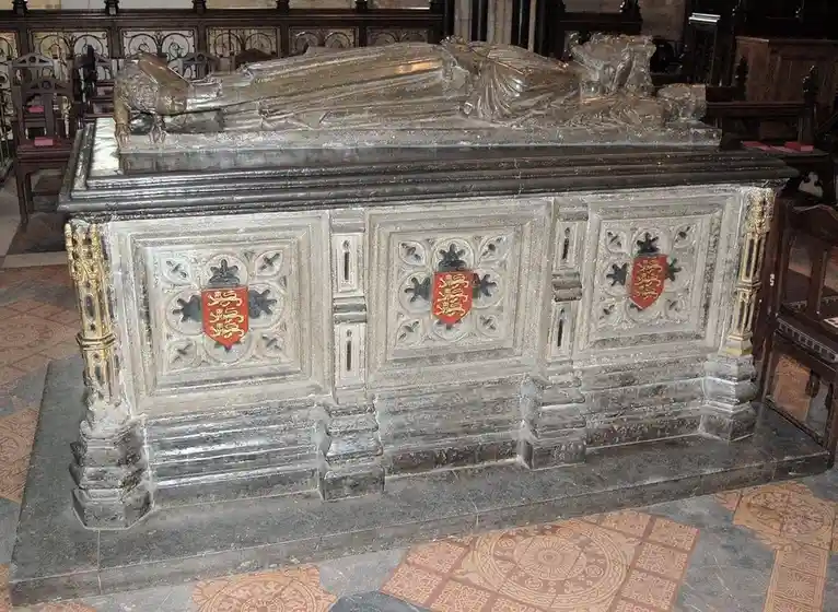 A large, ornate stone tomb with a reclining effigy of a person, featuring three red heraldic shields on its front panel, set…