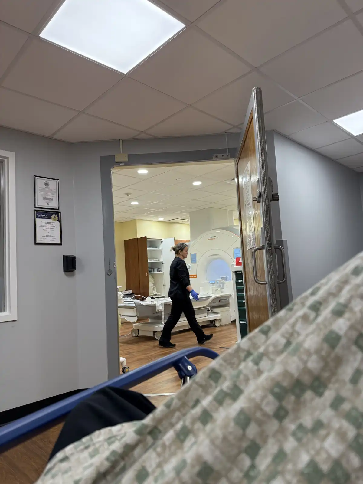A first-person perspective from a hospital bed looking toward an open door into a medical imaging room. A healthcare worker…