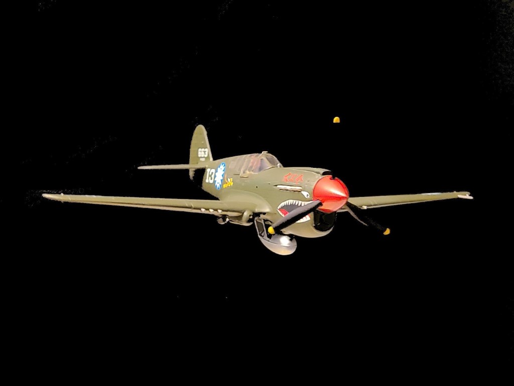 A green military fighter plane with a red nose cone and shark mouth painted on the front, flying against a black background…