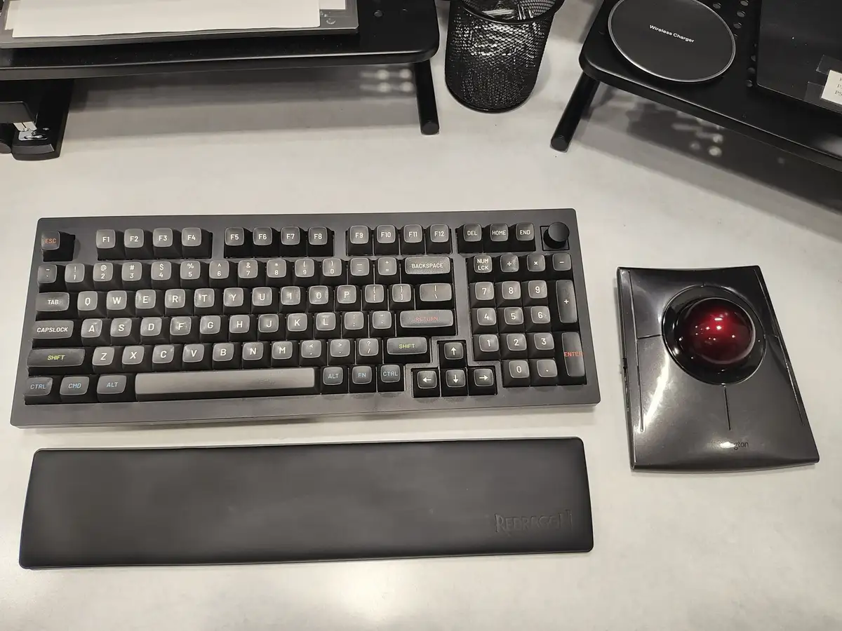 A black mechanical keyboard with gray and red keycaps sits on a white desk next to a black trackball mouse with a red ball…