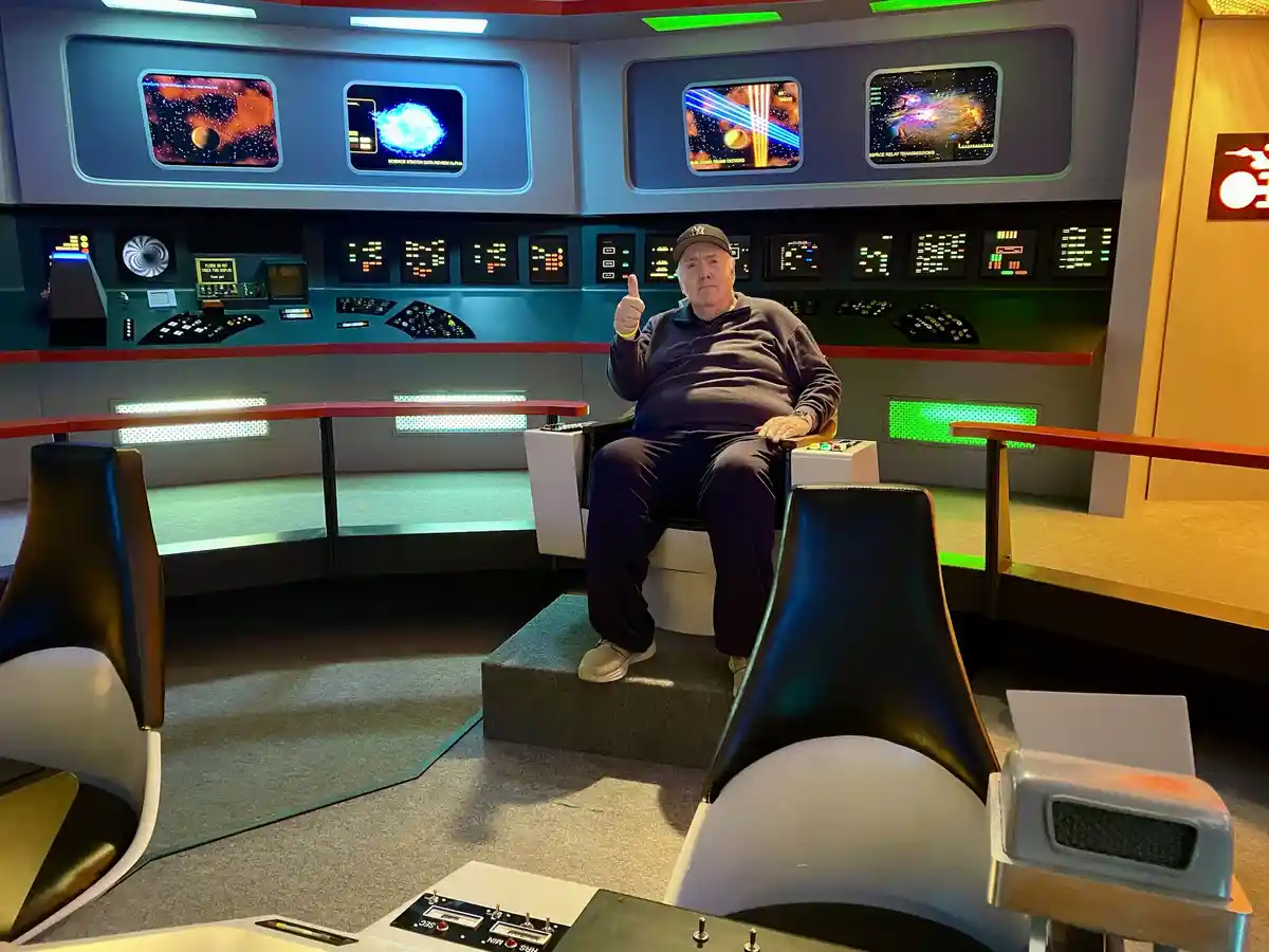 An older man wearing a baseball cap sits in a futuristic spaceship captain's chair, giving a thumbs-up. Behind him is a…
