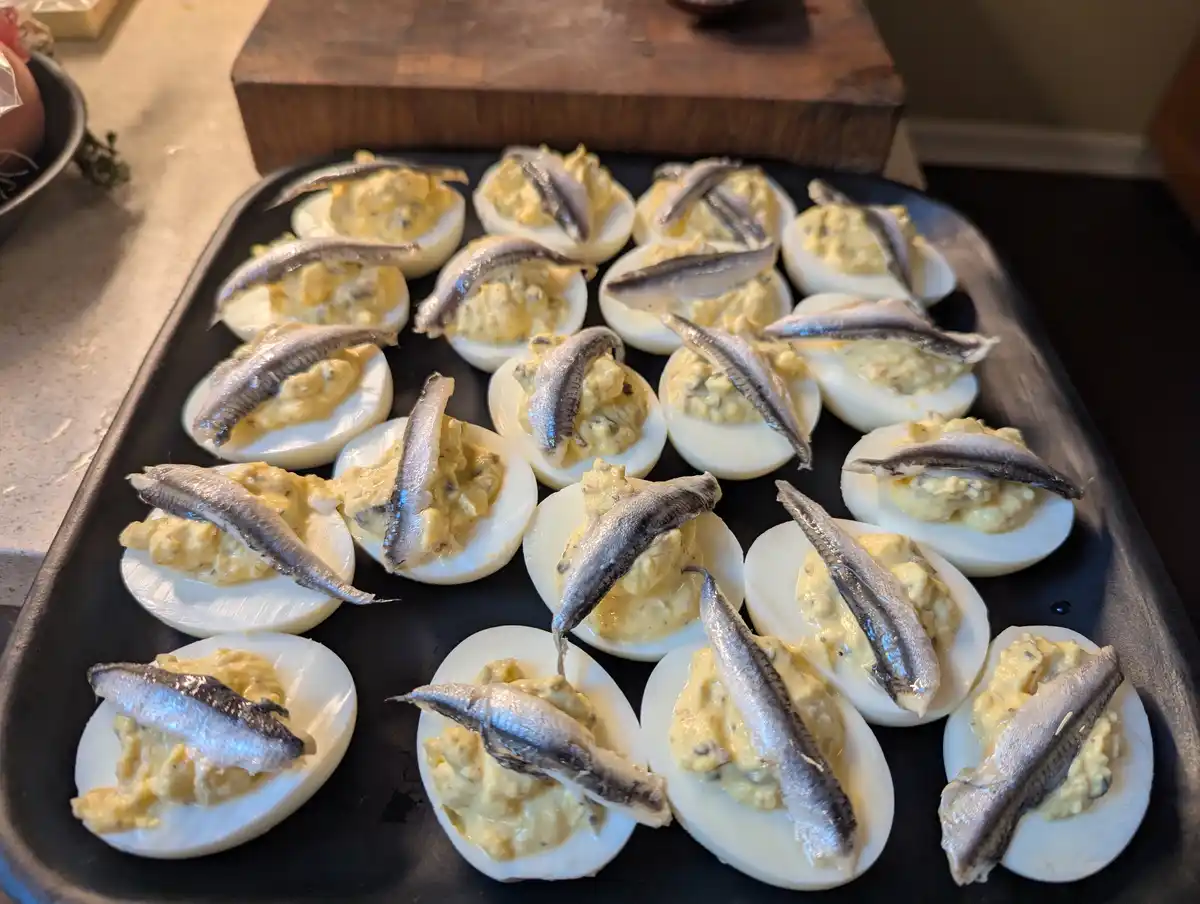 A black tray holds numerous deviled eggs, each topped with a small, whole anchovy. The eggs are arranged neatly on a kitchen…