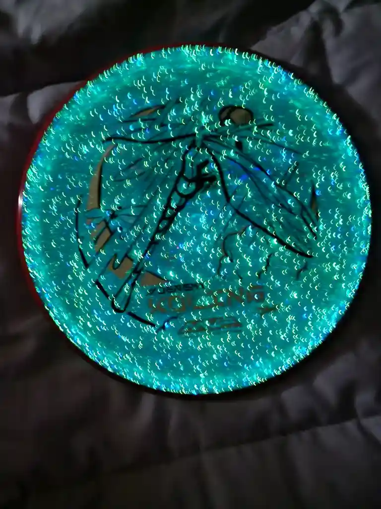 A close-up photo of a circular, iridescent green disc with a black outline of a dragonfly design, resting on a dark fabric…