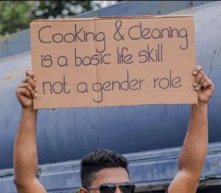 A person wearing sunglasses holds up a handmade cardboard sign with handwritten text stating 'Cooking & Cleaning is a basic…