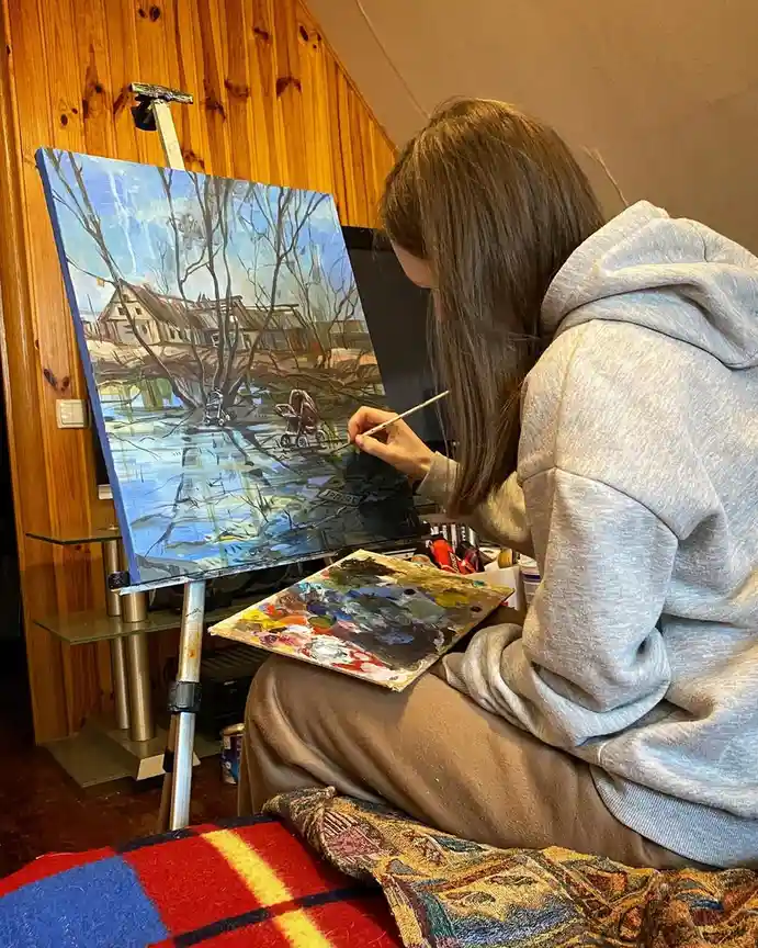 A person in a grey hoodie is painting a landscape on a canvas, with a stroller and a baby visible in the painting. The…