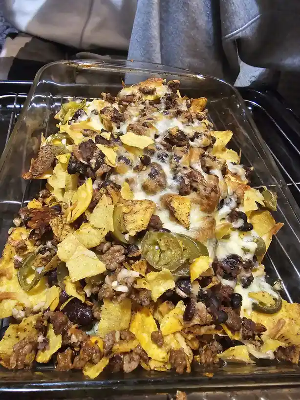 A close-up photo shows a glass baking dish filled with baked nachos, featuring melted cheese, ground meat, black beans, and…