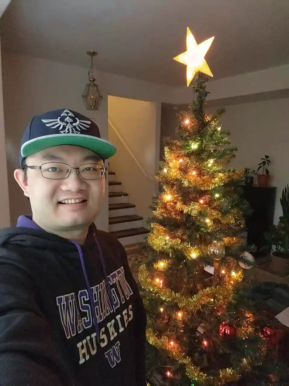 A smiling man wearing glasses, a blue and green baseball cap with a Zelda logo, and a black 'Washington Huskies' hoodie…