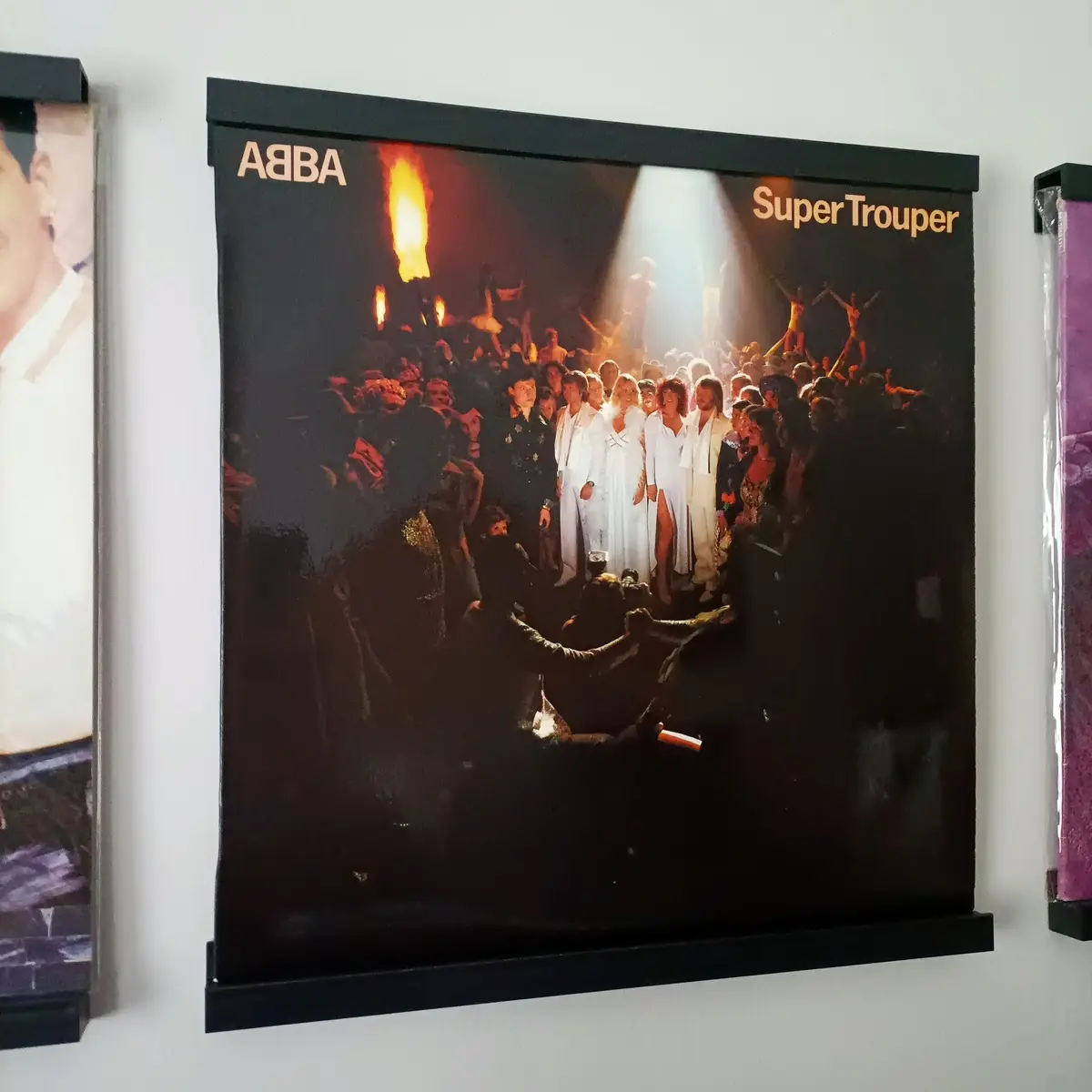 A framed poster of the ABBA album 'Super Trouper' is mounted on a wall, showing the band in white outfits on stage with a…