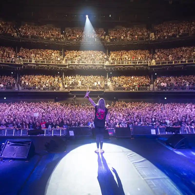 A musician stands alone on a brightly lit stage, facing a massive, multi-tiered arena crowd with arms raised. The audience…