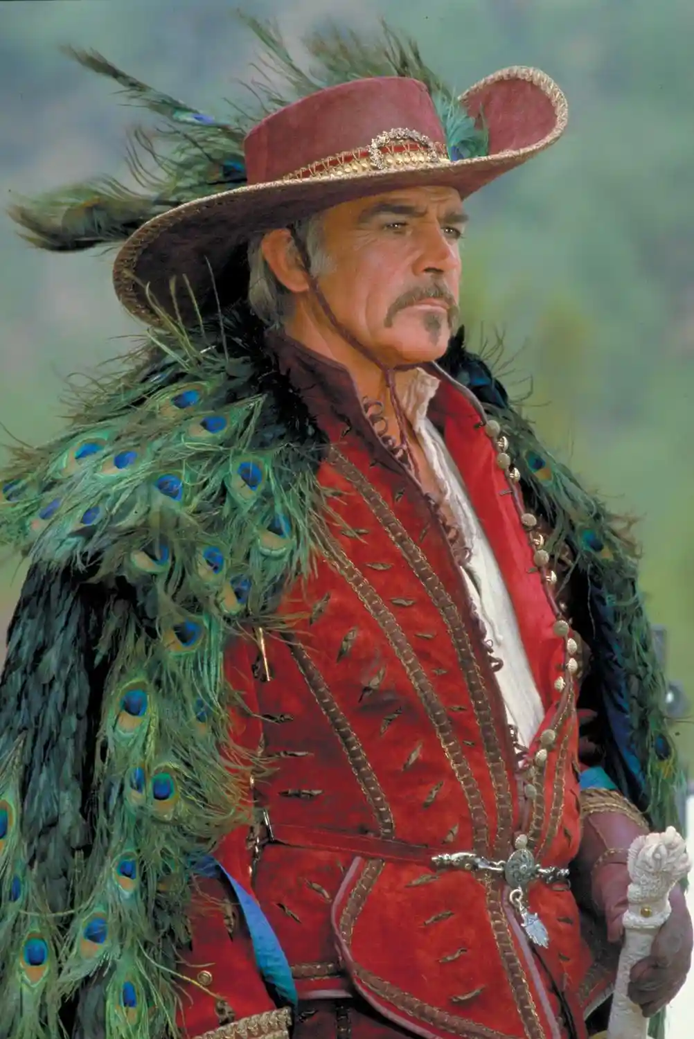 A man in a red velvet jacket and a wide-brimmed hat adorned with peacock feathers looks off to the side. He wears a matching…