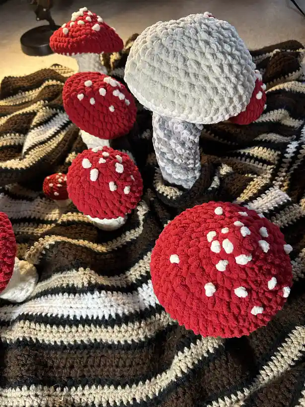 Several crocheted mushrooms, mostly red with white spots like fly agarics, are arranged on a striped crocheted blanket. One…