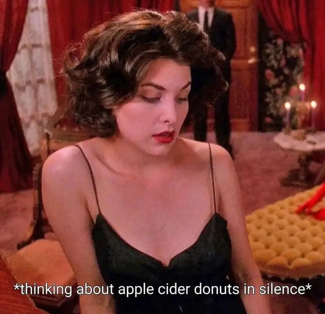 A woman with dark curly hair and red lipstick, wearing a black spaghetti strap top, looks down with a pensive expression in…