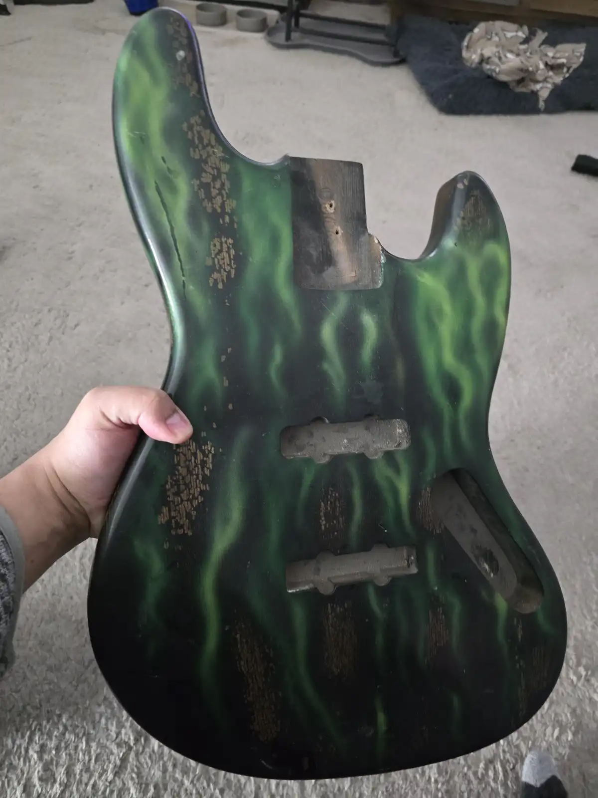 A hand holds up the back of a guitar body with a green and black flame finish, showing significant wear and tear, including…