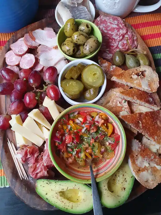 An overhead photo of a wooden board laden with various foods including sliced meats, cheese, grapes, olives, pickles…