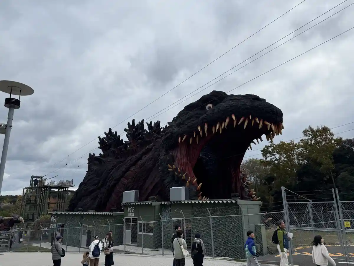 A large, realistic Godzilla statue with an open mouth full of sharp teeth and a row of dorsal fins stands behind a fence…