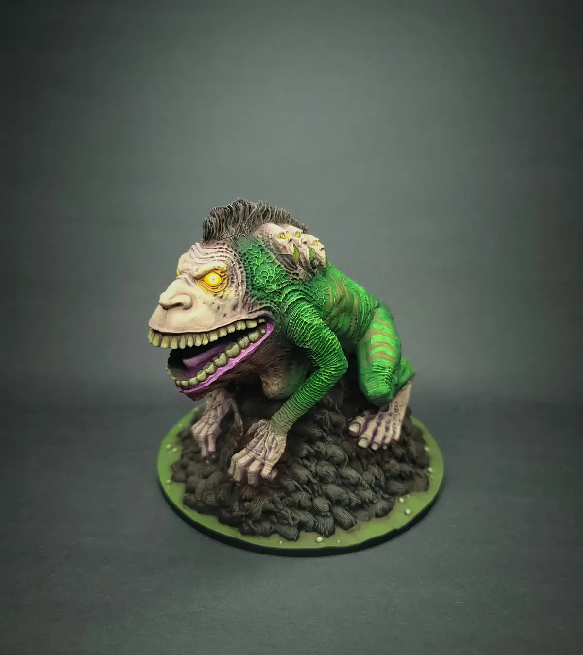 A painted miniature monster figurine, resembling a frog with a human-like face and a mohawk hairstyle, crouches on a rocky…