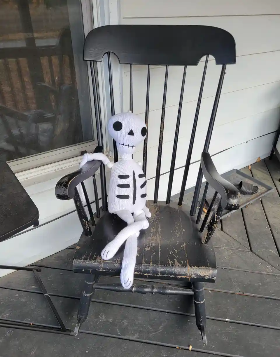 A white knitted skeleton plush toy sits in a black, weathered rocking chair on a porch, with one arm resting on the chair's…