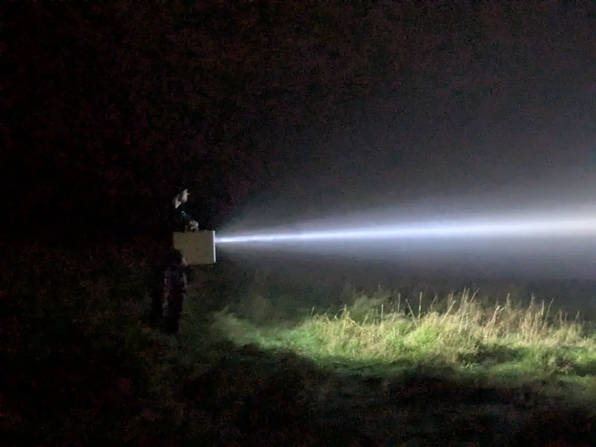 A person stands in a dark field at night, holding a large, boxy flashlight that emits an extremely bright, wide beam of…