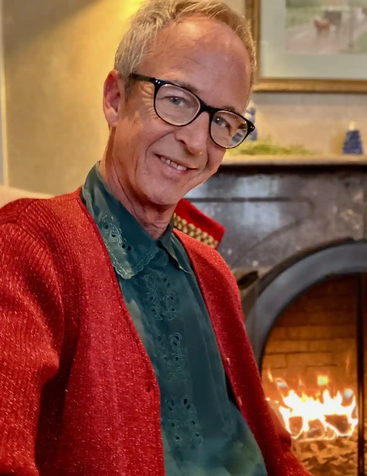 A middle-aged man with glasses smiles at the camera while wearing a sparkly red cardigan over a teal embroidered shirt…
