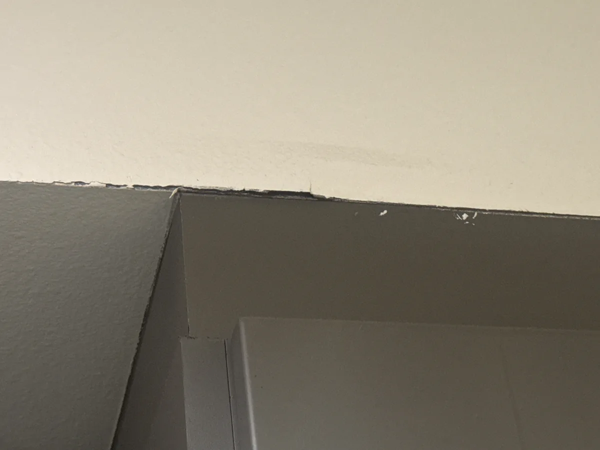 A close-up photo shows the corner where a dark gray painted wall meets a white ceiling, with visible cracks and peeling…