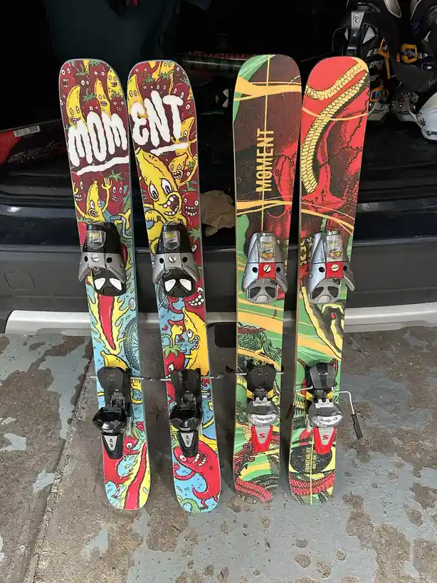 Two pairs of colorful, cartoonishly designed Moment brand skis stand upright on a concrete floor, leaning against the open…
