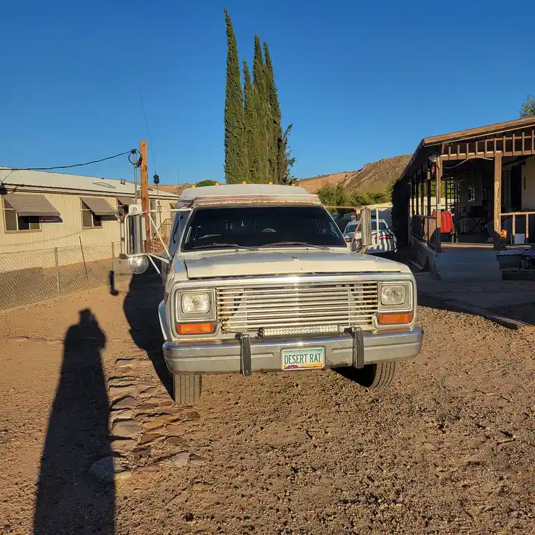 A white, older model SUV is parked on gravel in a desert-like residential area with a clear blue sky, a tall cypress tree…