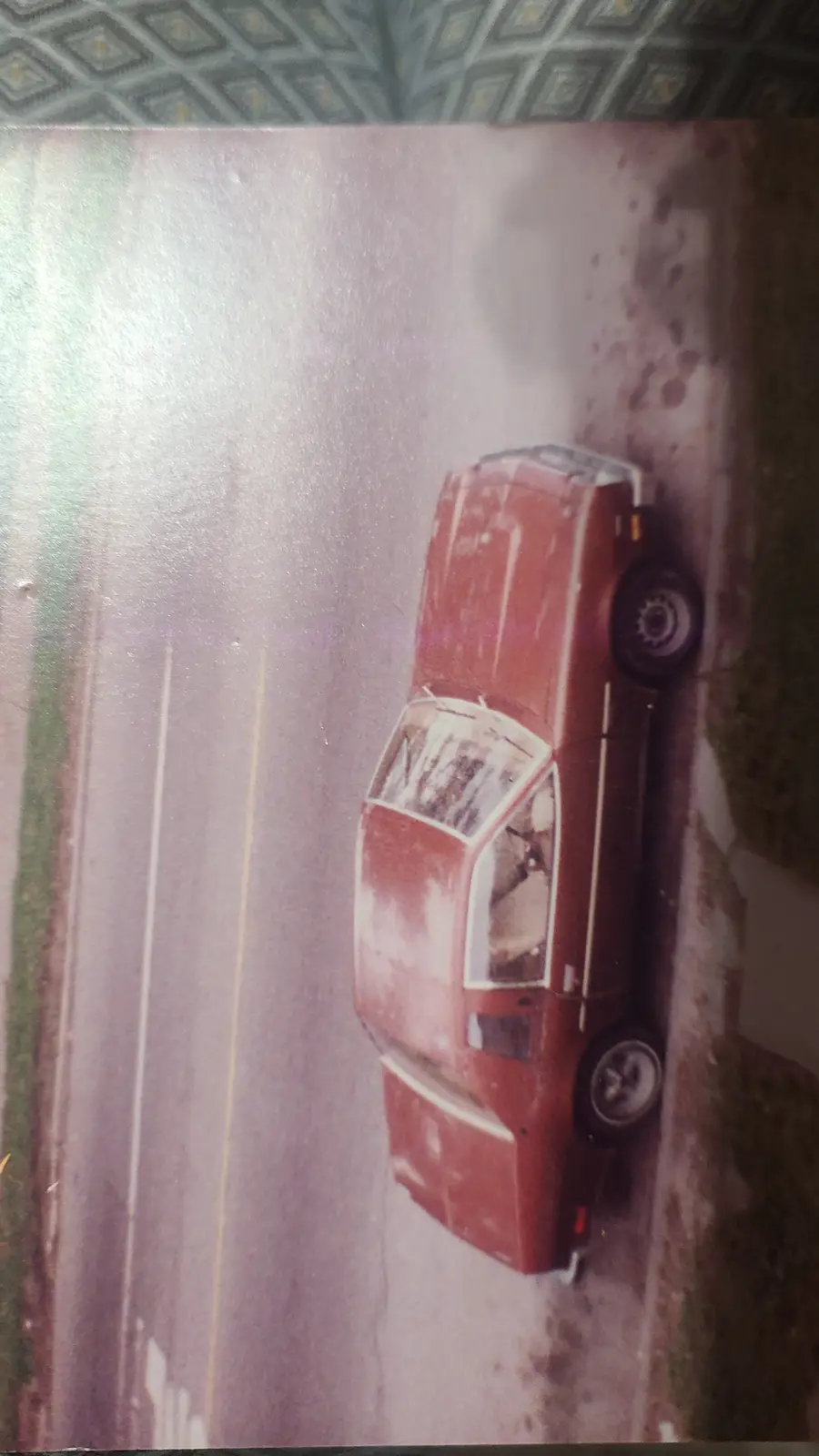 A vintage red car is shown upside down on a road, with its wheels pointing skyward and the roof touching the ground. The…