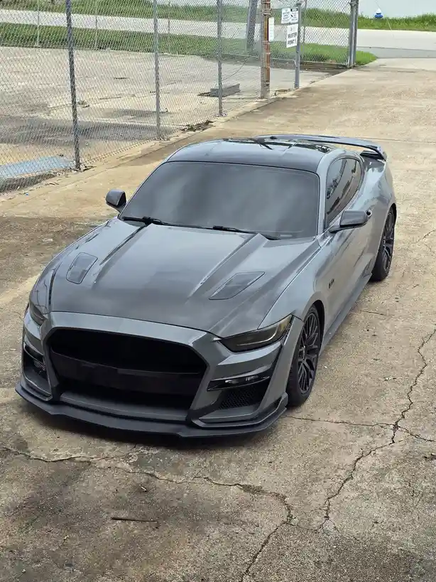 A matte gray Ford Mustang with a black hood and aggressive front bumper sits parked on cracked concrete near a chain-link…