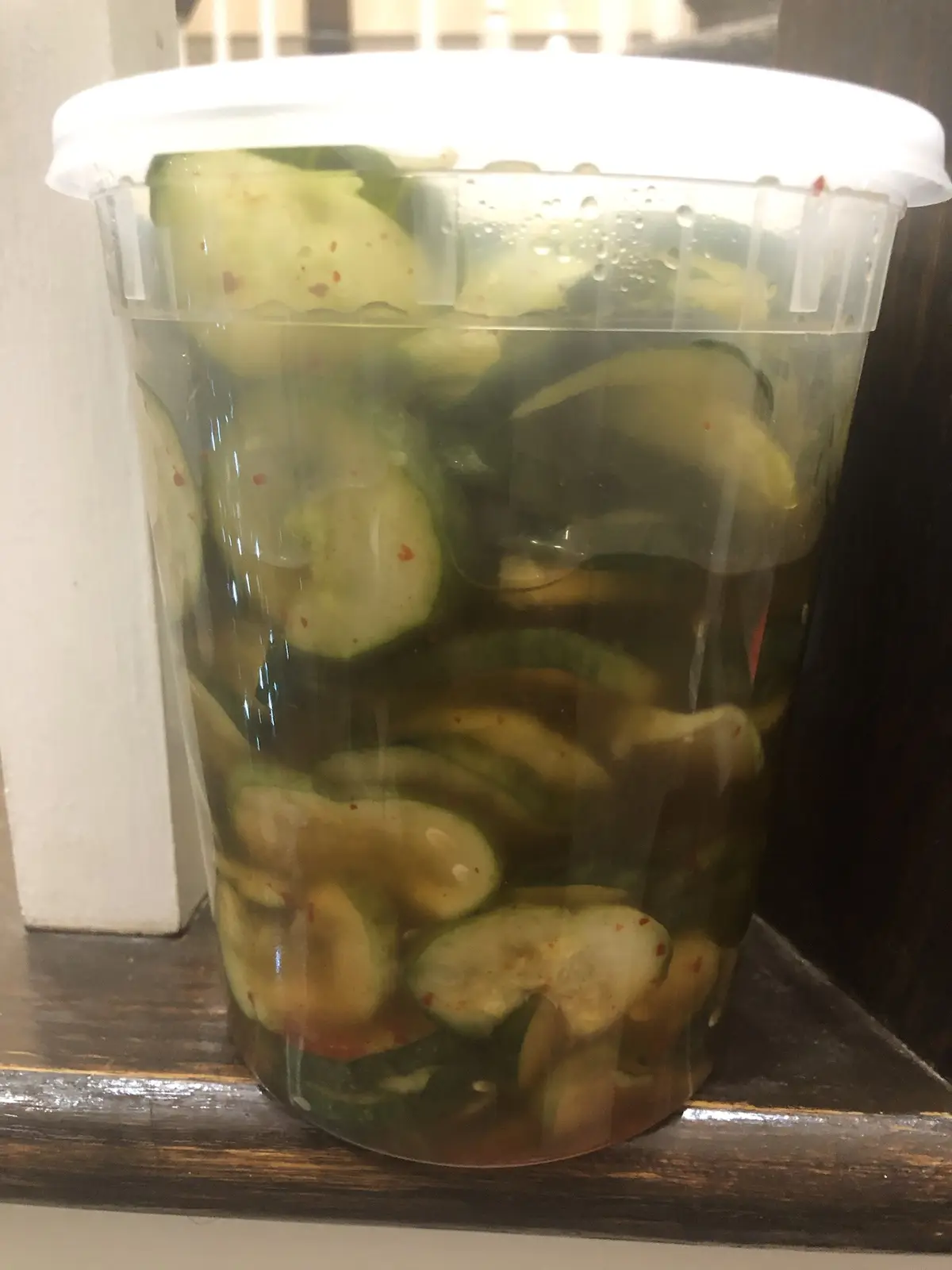 A clear plastic container with a white lid is filled with sliced cucumbers submerged in brine, showing condensation on the…
