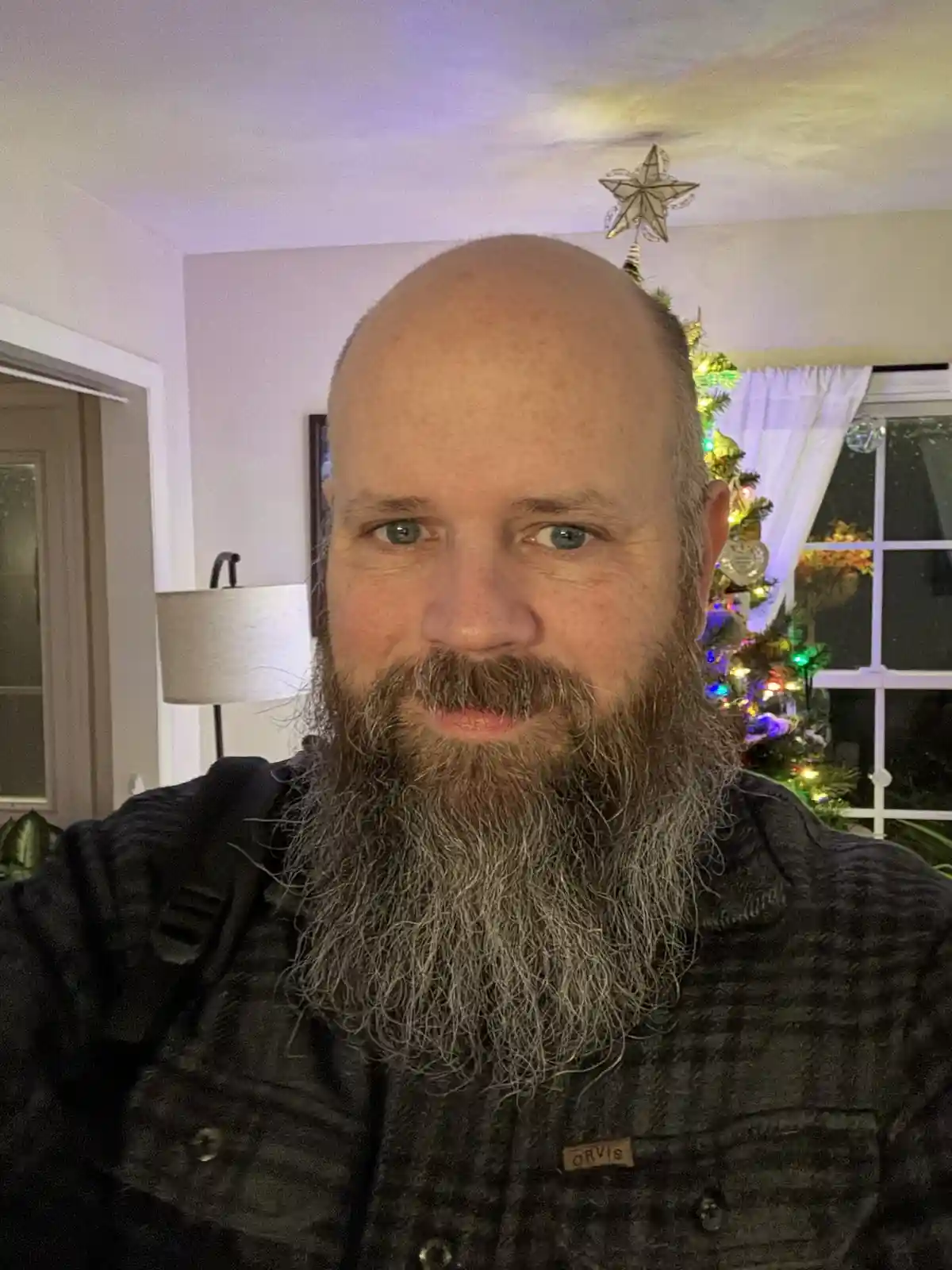A man with a full, graying beard and a bald head looks directly at the camera, wearing a dark jacket with an 'ORVIS' patch…