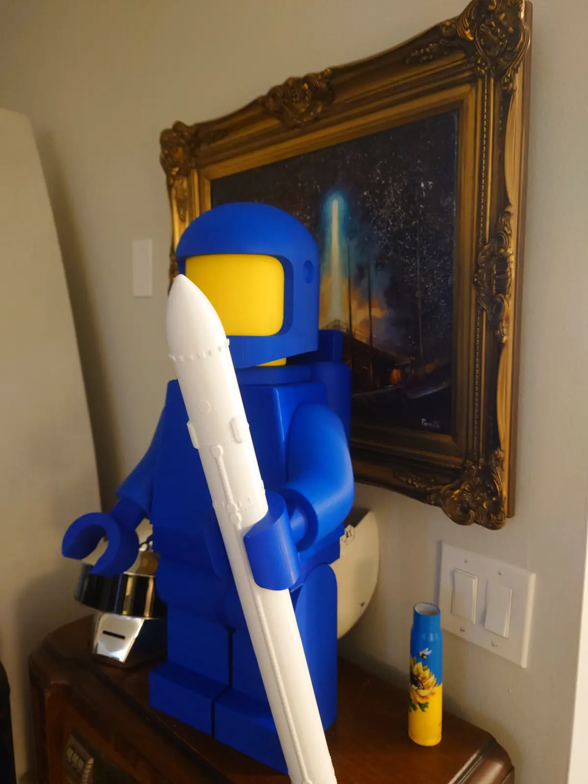 A large blue LEGO astronaut figure holding a white rocket stands on a wooden table in front of a framed painting depicting a…