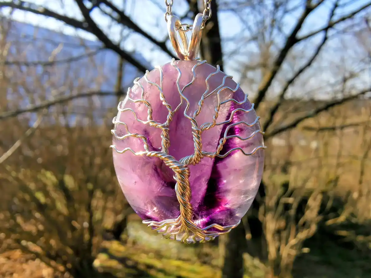 A close-up photo shows a pendant made of a round, translucent purple amethyst stone, intricately wrapped with silver and…