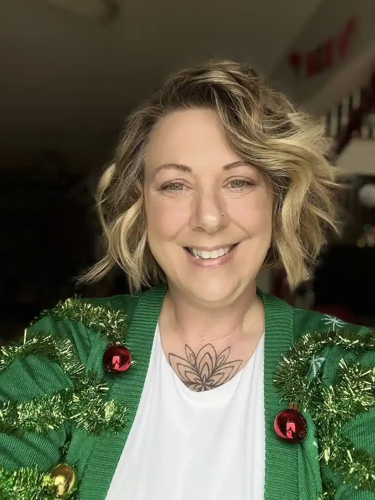 A smiling woman with short blonde hair wears a green cardigan decorated with tinsel and red ornaments, resembling a…