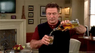 A man in a red shirt and black vest pours amber liquid from a bottle into a glass, his expression showing reluctance or…