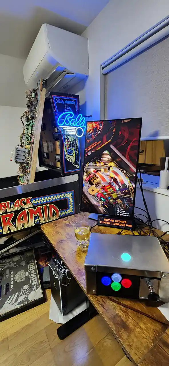 A custom-built pinball machine labeled 'BLACK PYRAMID' with its internal wiring exposed, sits next to a monitor displaying a…