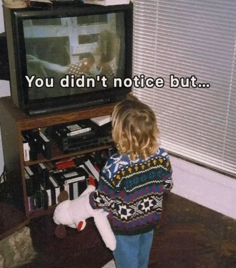 A young child with curly hair, wearing a colorful patterned sweater and blue jeans, stands in front of an old CRT…